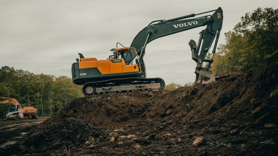 Tracked excavator shaping a graded dirt building pad on a wooded lot